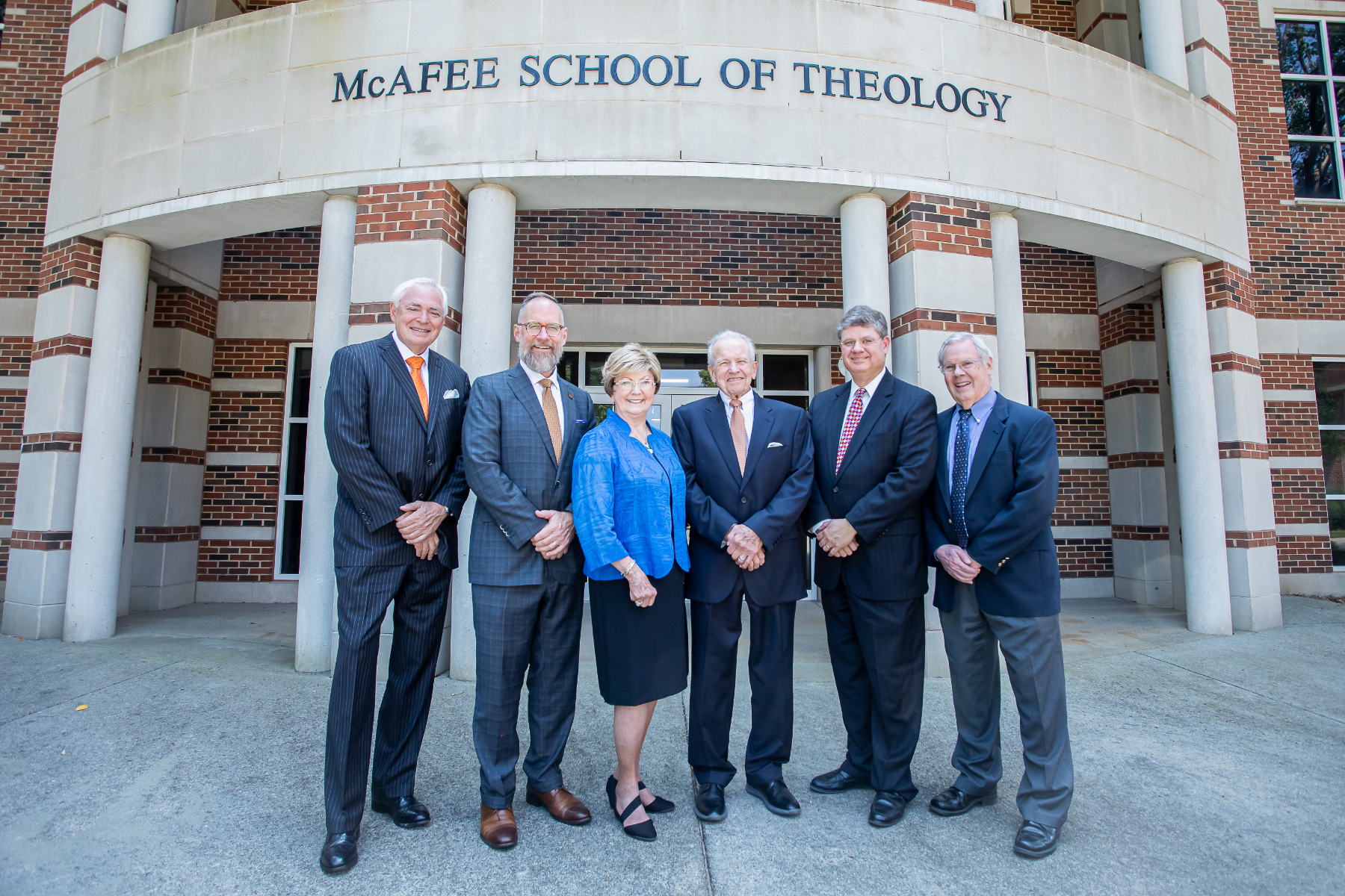 Six people wearing business attire stand in front of a brick building identified as McAfee School of Theology.