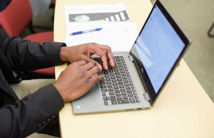 Person working on a laptop with documents on a desk in a classroom setting.