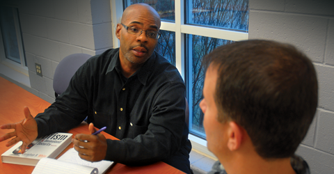 Two individuals engaged in a discussion at a table with a book, in a room with a window showing trees outside.