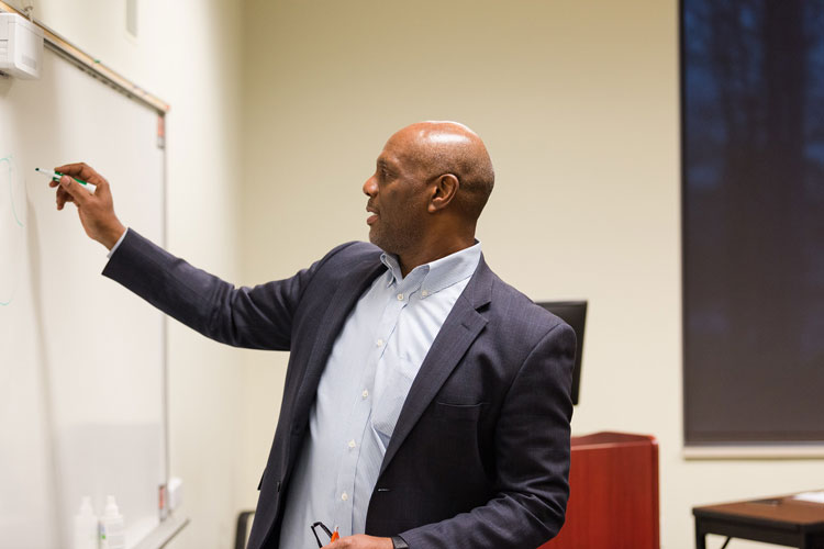 A man wearing a collared shirt and blazer writes on a white board in a classroom setting.