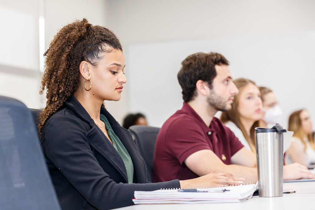 Adults seated in a classroom setting, listening attentively. One person in the foreground is taking notes beside a metal water bottle.