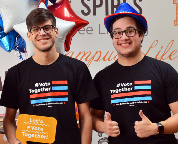 Two students give thumbs up while wear black T-shirts that say "Vote Together." One is holding an orange sign that says "Let's Vote Together."