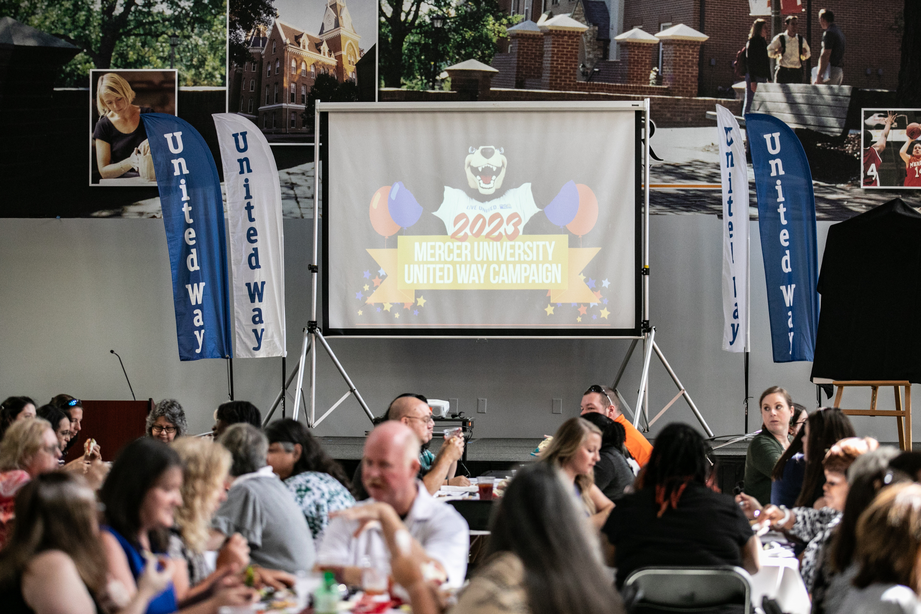 A projector screen showing an image of Toby and a banner that says 2023 Mercer University United Way Campaign. People can be seen eating at tables.