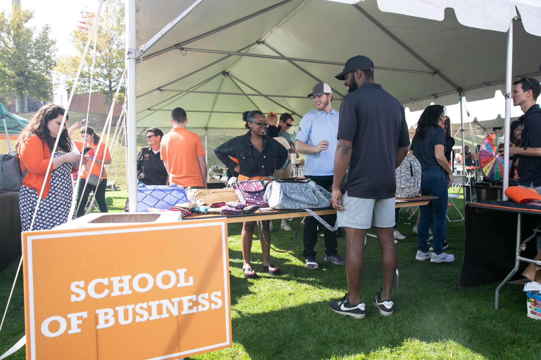 Tables set up under a large white tent outdoors, with people milling around. A sign in the grass says, "School of Business."