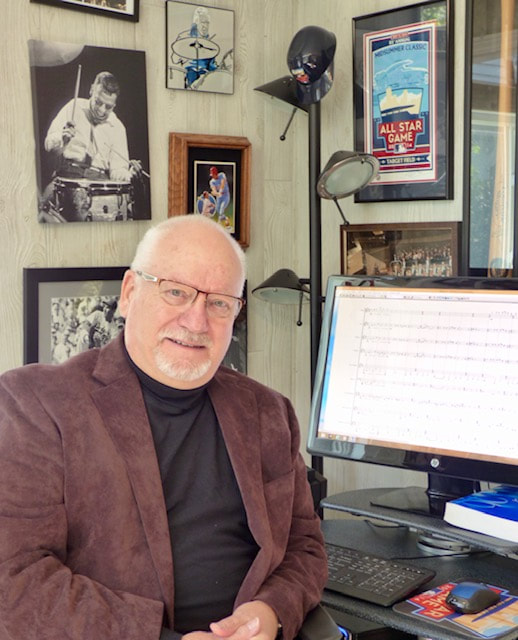 A man in a black turtleneck shirt and brown blazer sits next to a computer monitor displaying staff music.