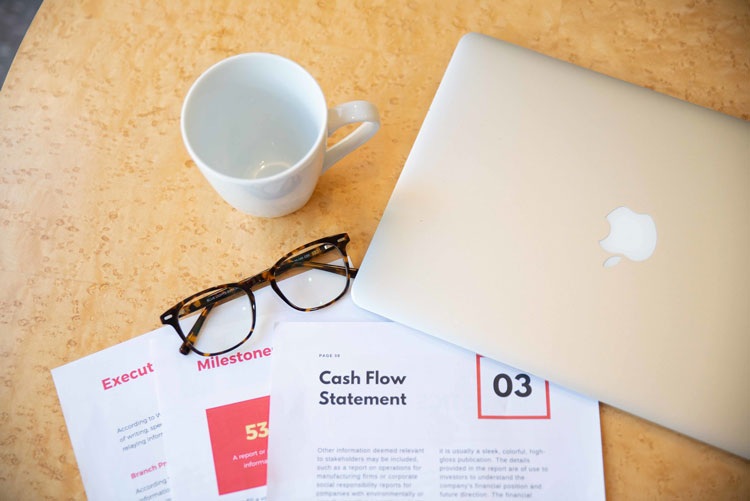 A workspace featuring a closed laptop, a coffee mug, eyeglasses, and documents with labels like "Cash Flow Statement" on a textured table.