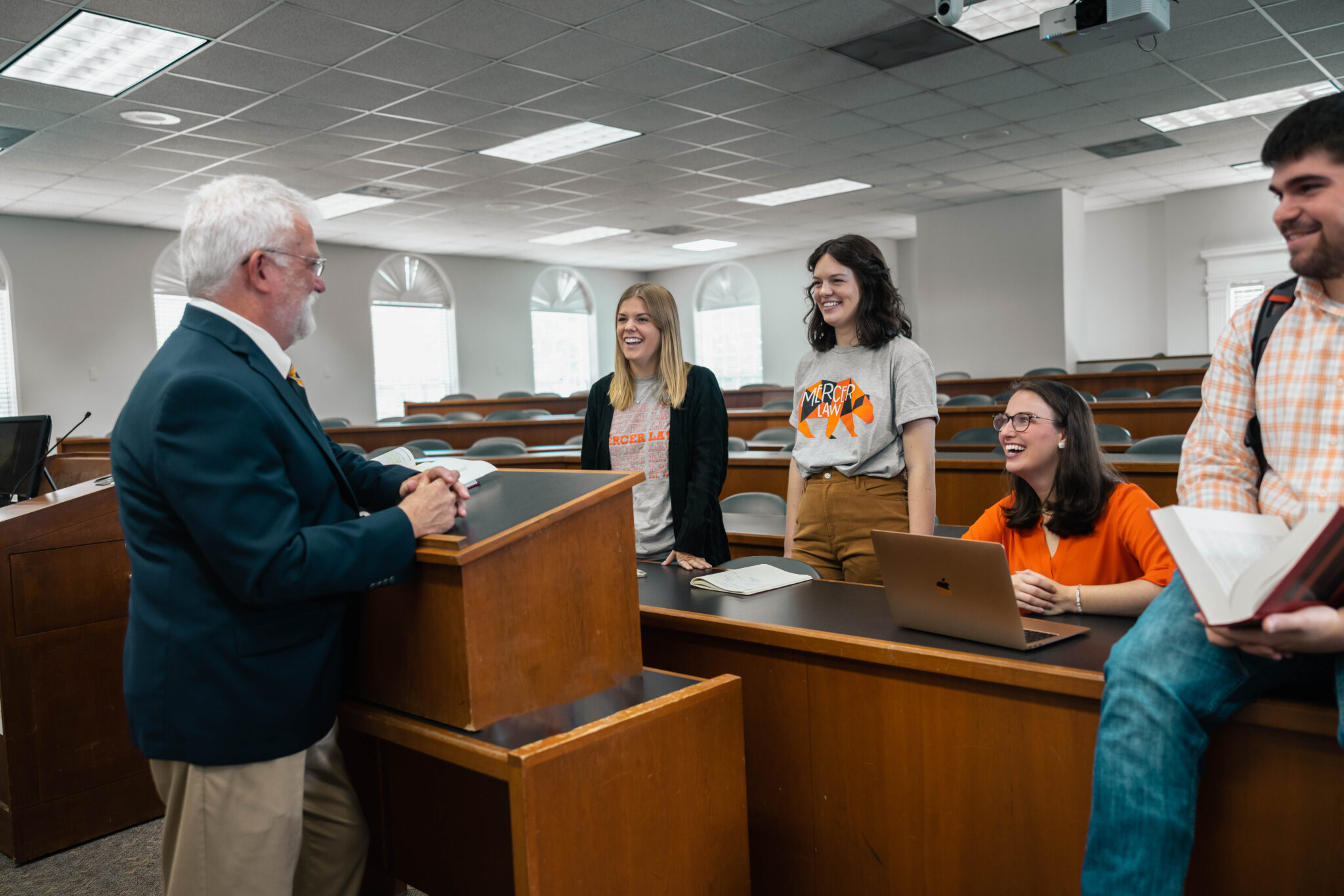 Four people sit in the front row of a classroom interacting with a professor behind a podium.