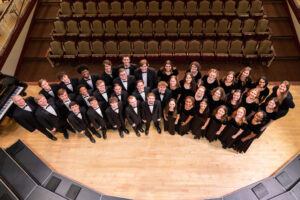 Men and women wearing black tuxedos and dresses stand in a semicircle on stage.