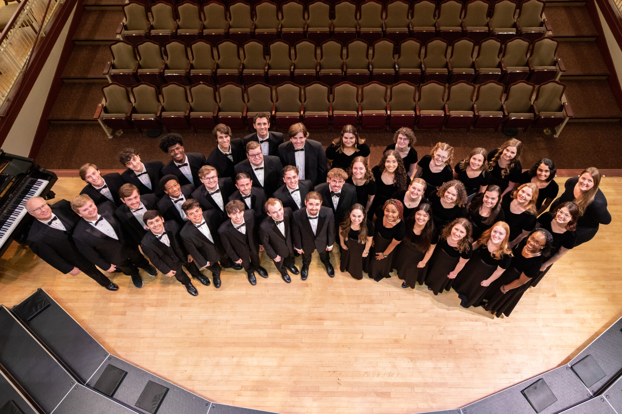 Men and women wearing black tuxedos and dresses stand in a semicircle on stage.