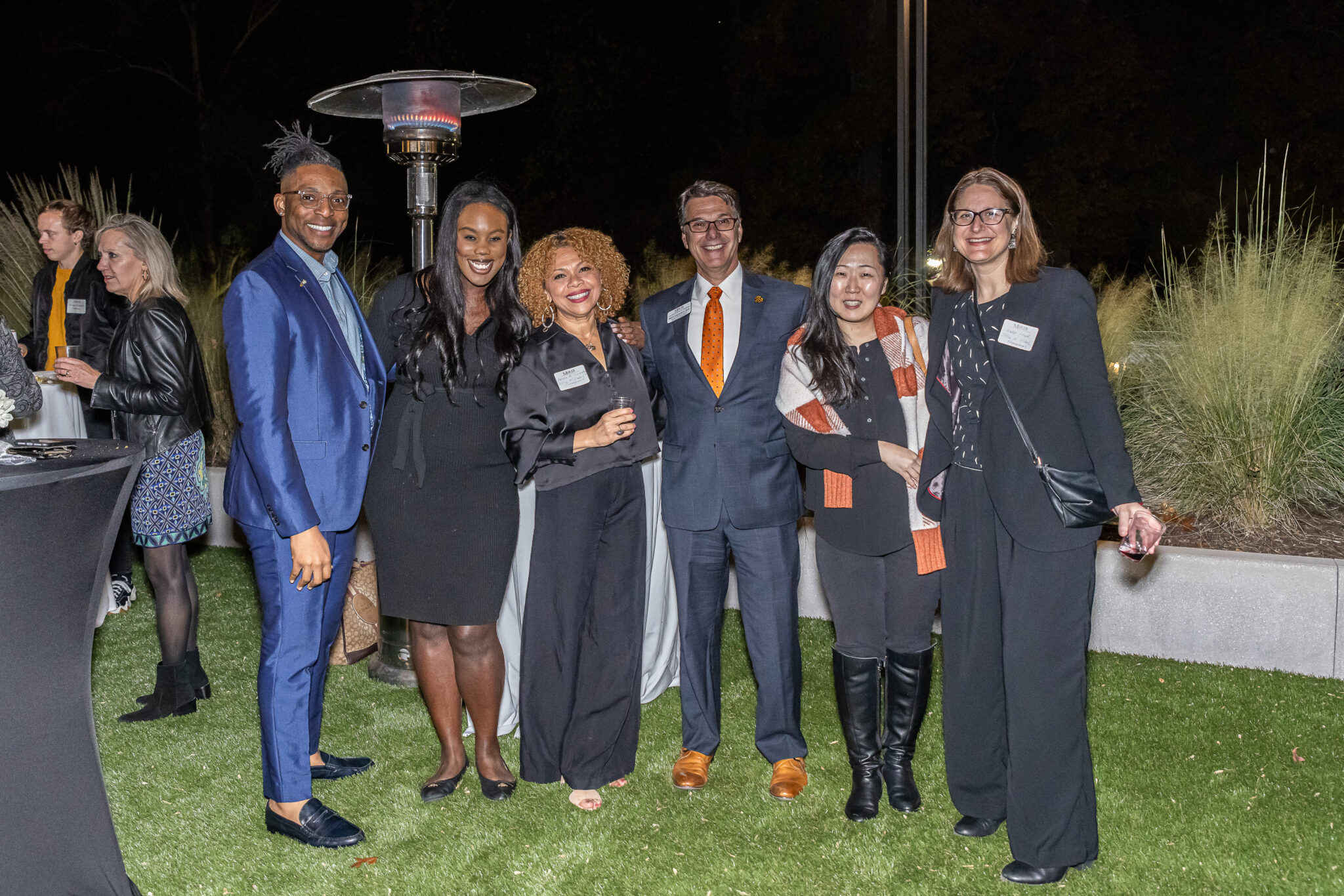 A group of six individuals in cocktail attire pose for a photo in a grassy area while others mingle in the background.