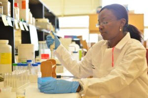 A Mercer student in a lab coat and gloves uses a pipette to transfer liquid into a test tube. The laboratory is filled with various bottles and lab equipment.