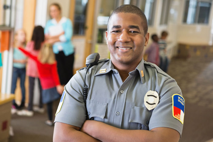 Public safety officer smiling at the camera in a school hallway, wearing a uniform with a badge. In the background, a teacher interacts with children.