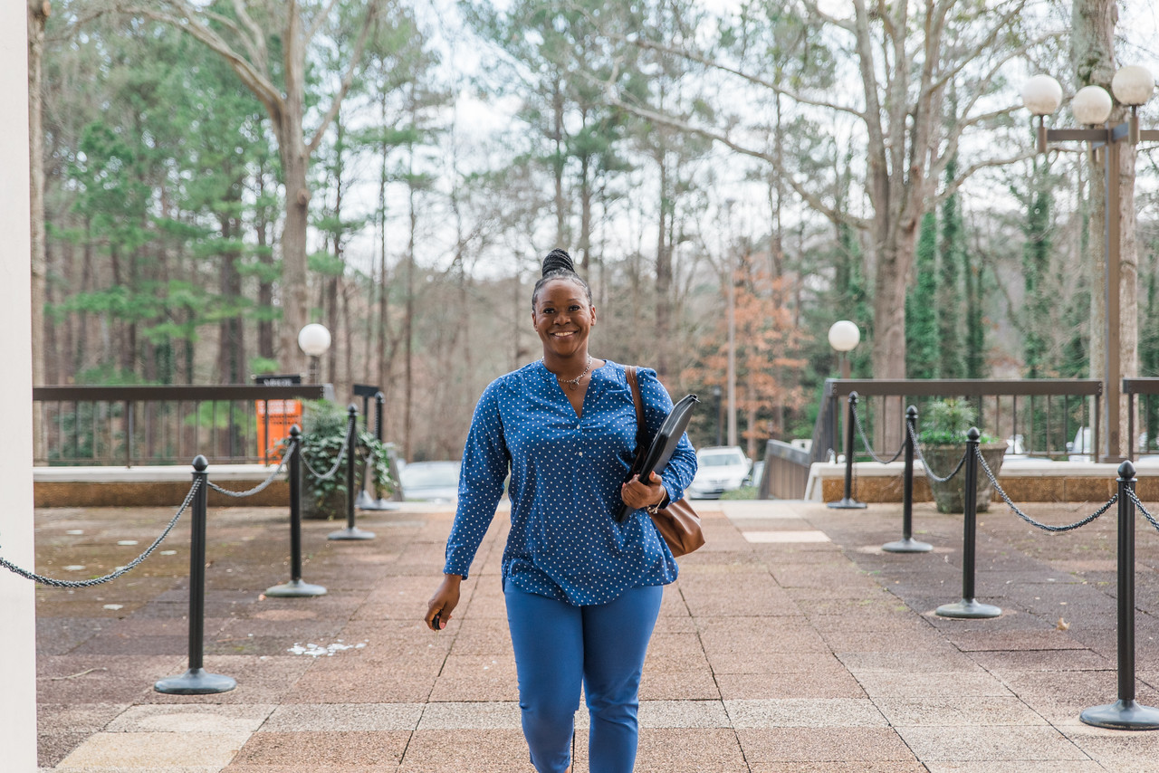 Smiling person walking on a university campus path with trees and lamp posts in the background, carrying a bag and a zipper binder.