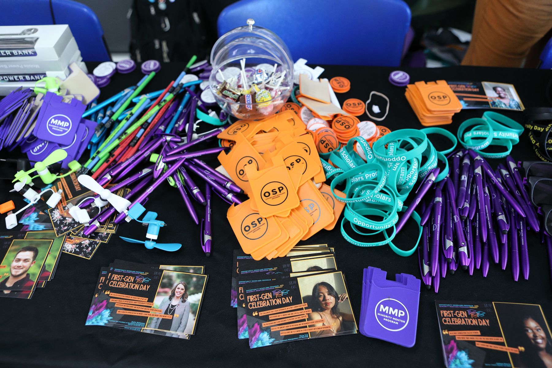 Freebies on a table, including pens, candy, cellphone wallets, and rubber bracelets. Postcards on the table say, "First-Gen Celebration Day."