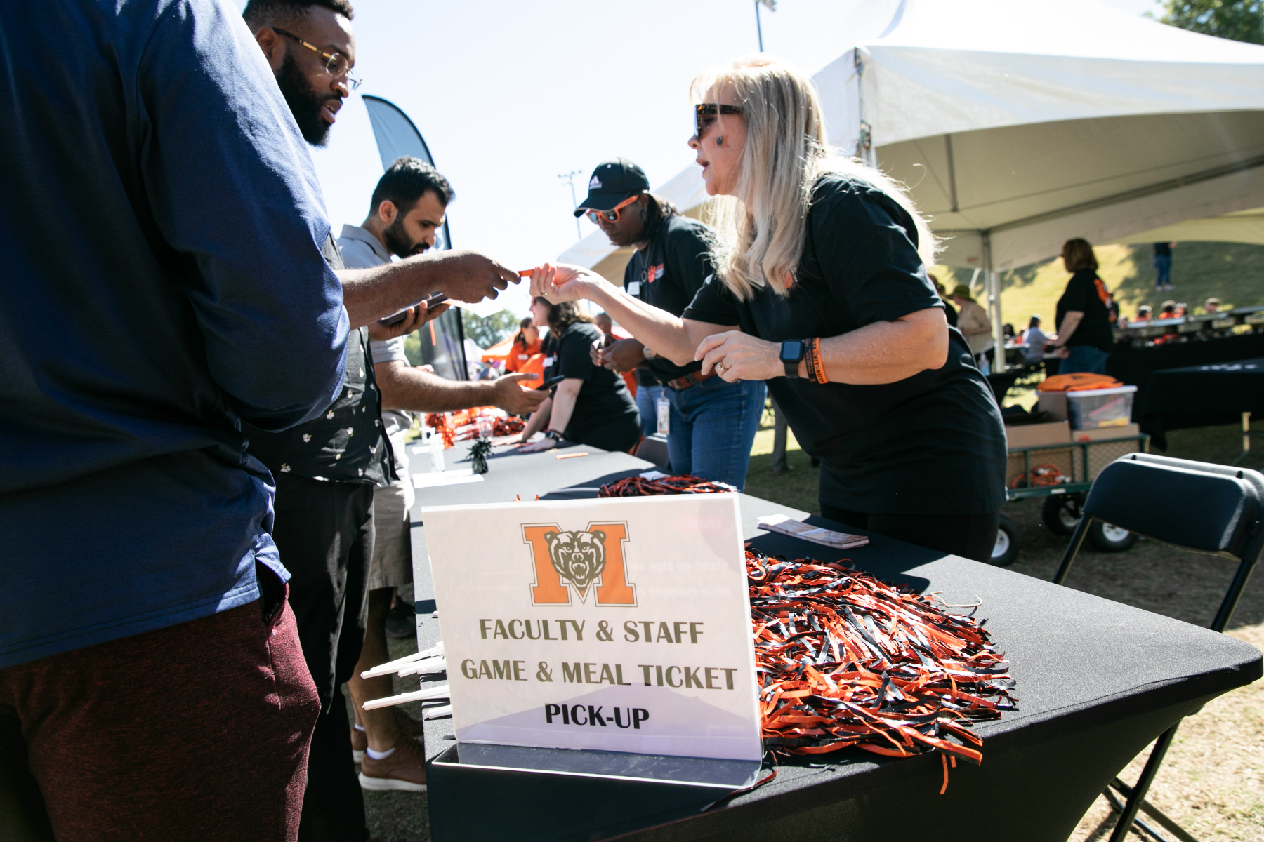 People gather at a table with a sign reading "FACULTY & STAFF GAME & MEAL TICKET PICK-UP" at an outdoor event, exchanging tickets and items.