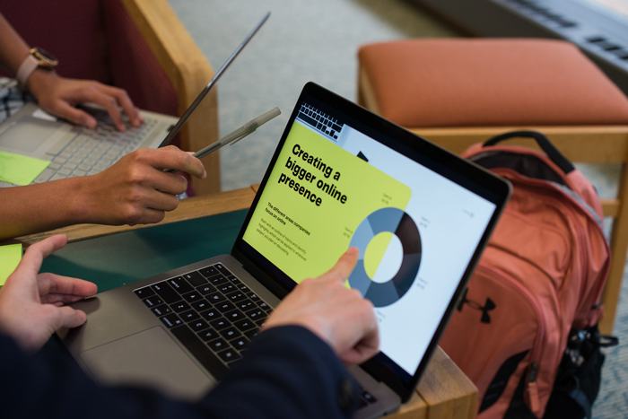 Two individuals collaborating at a table with laptops; one laptop screen displays a presentation on creating a bigger online presence.