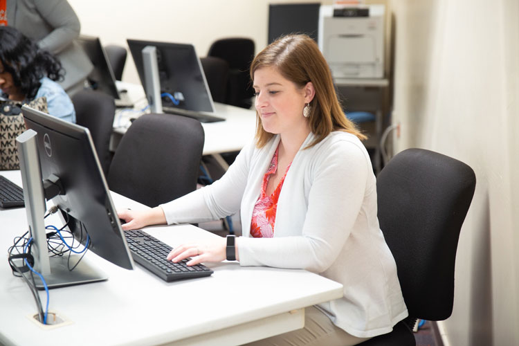 A person is seated at a desk in a computer lab, working intently on a desktop computer. They are wearing a white cardigan over an orange blouse. There are other individuals in the background, also engaged with computers. The room is well-lit and equipped with multiple workstations.