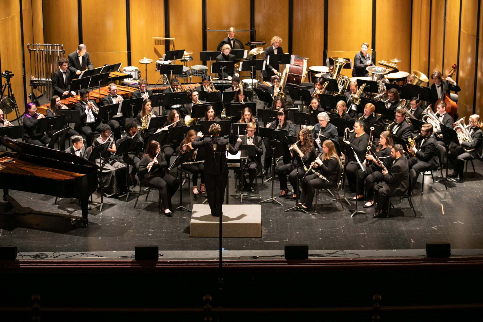 A band performs on stage, featuring various musicians with instruments such as brass, woodwinds, percussion and a grand piano.