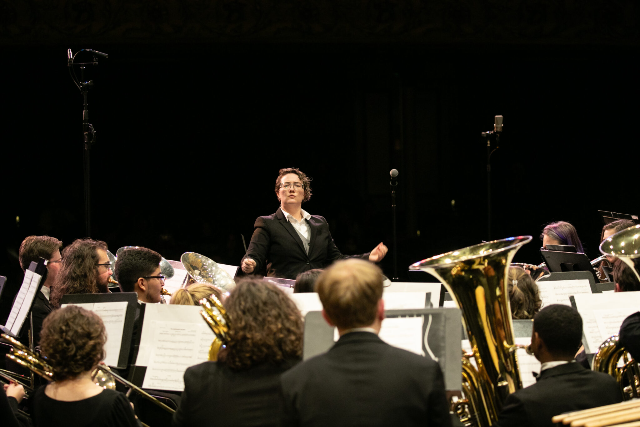 Conductor leading a wind ensemble performance on stage, viewed from behind the brass section.