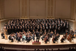 A large choir and orchestra performing onstage at Carnegie Hall, conducted by a central figure in formal attire.