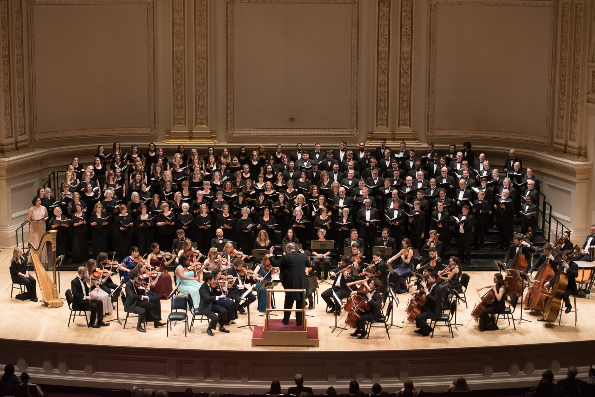 A large choir and orchestra performing onstage at Carnegie Hall, conducted by a central figure in formal attire.