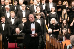 A conductor in a black suit and bow tie leading a choral and orchestral performance in a concert hall, with performers in black attire visible in the background.