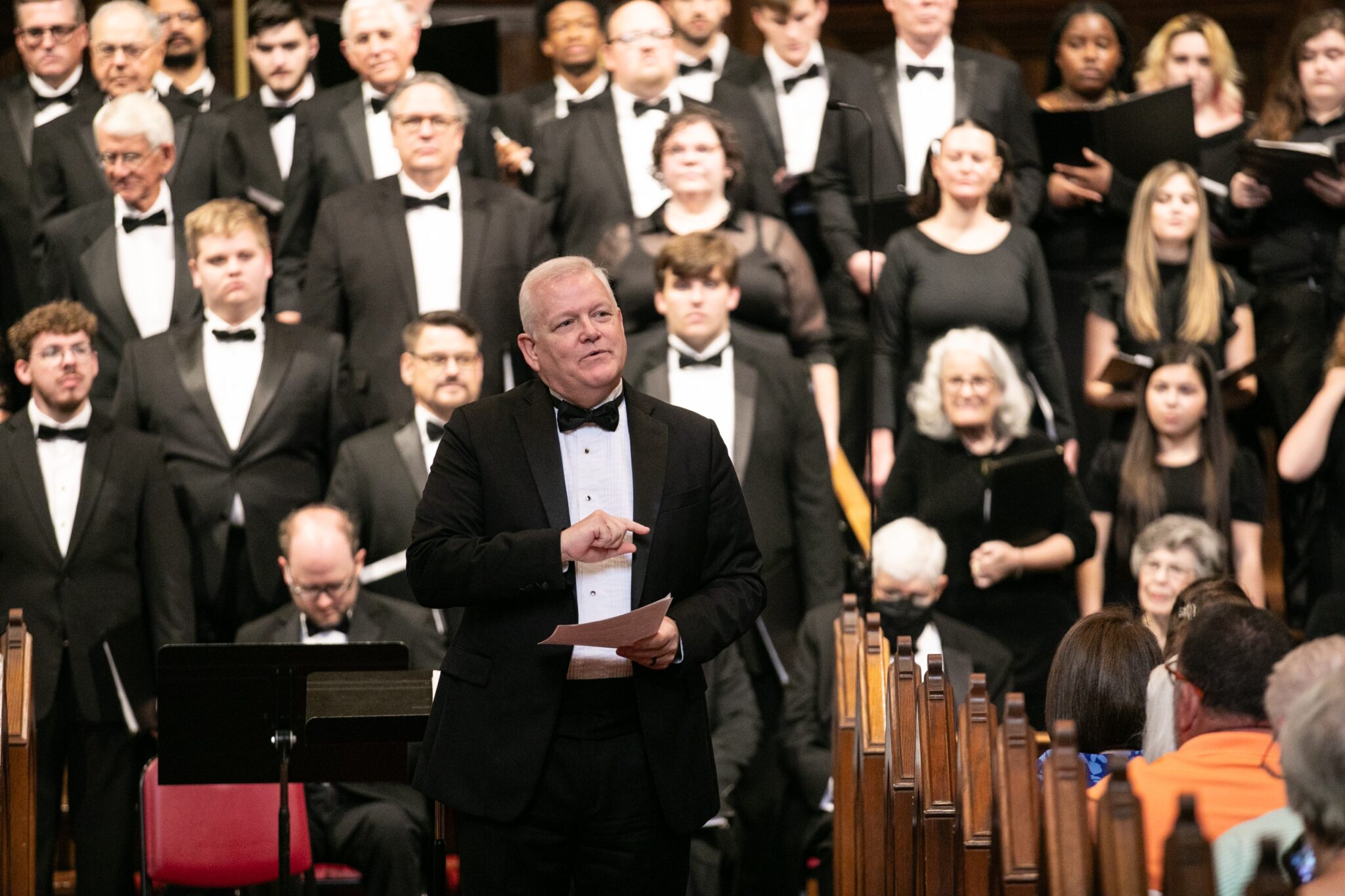 A conductor in a black suit and bow tie leading a choral and orchestral performance in a concert hall, with performers in black attire visible in the background.