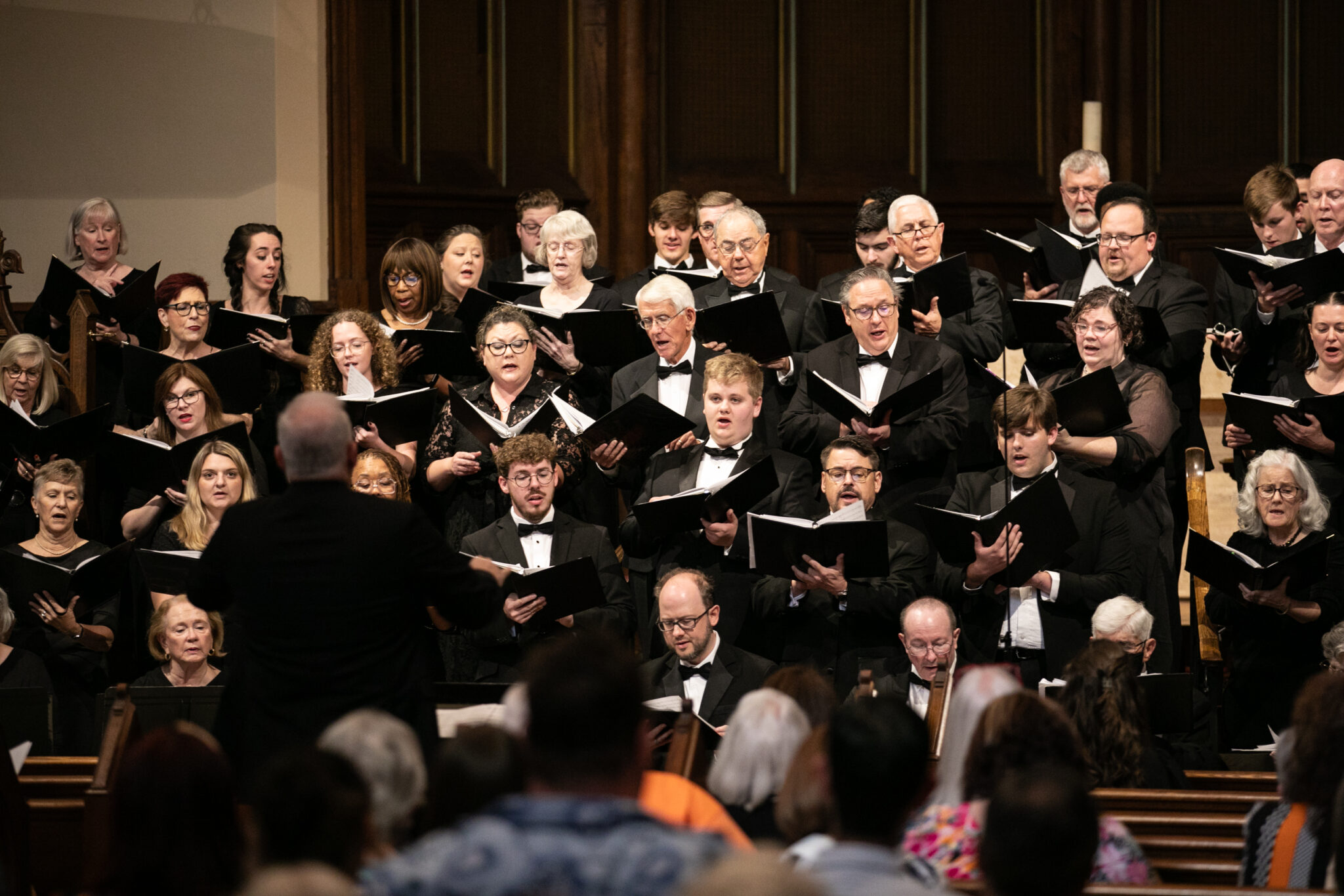 A choir performing on stage, with a conductor directing them, in front of an audience.