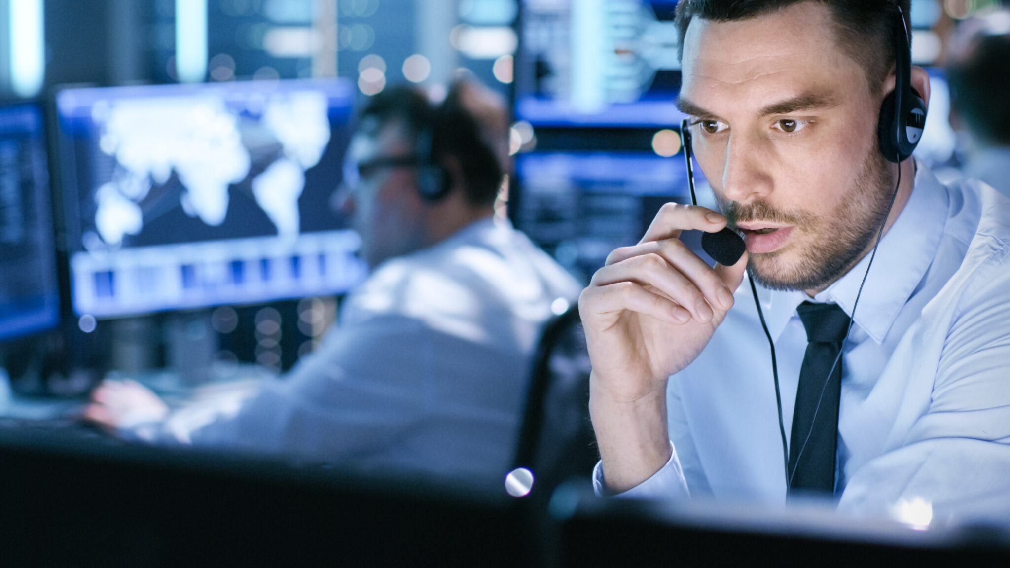 A person using a headset with a microphone in a busy control room environment, focused intently on multiple computer monitors. In the background, another individual is also looking at a computer screen. The setting suggests a high-tech or security monitoring center.