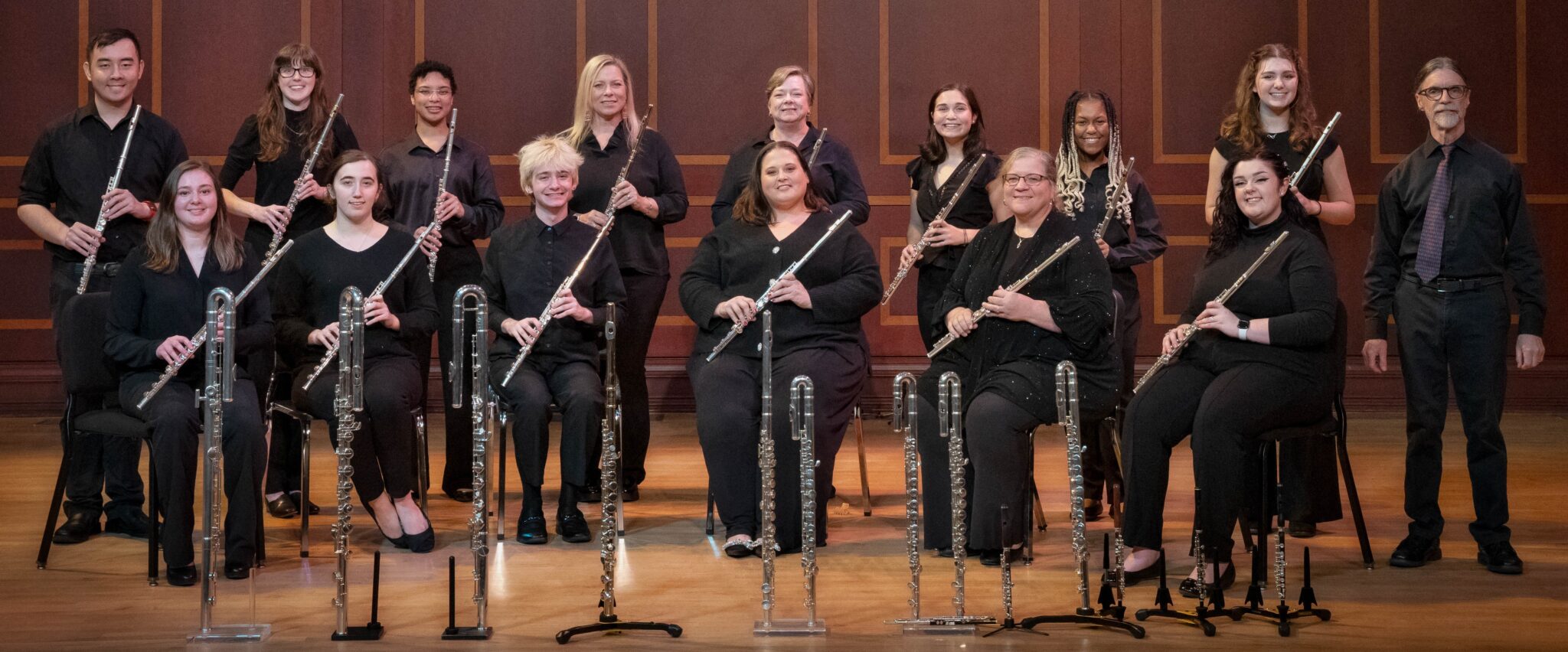 A group of 14 flutists wearing black attire, seated and standing in two rows on a stage, each holding a flute, in an elegant concert hall setting. A conductor stands to the right.