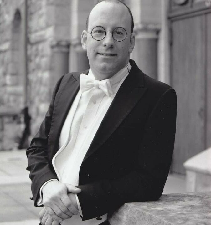 A portrait of an individual wearing a tuxedo with a bow tie, standing against a historical building. The person is smiling, wearing round glasses, and has their hands lightly clasped in front.