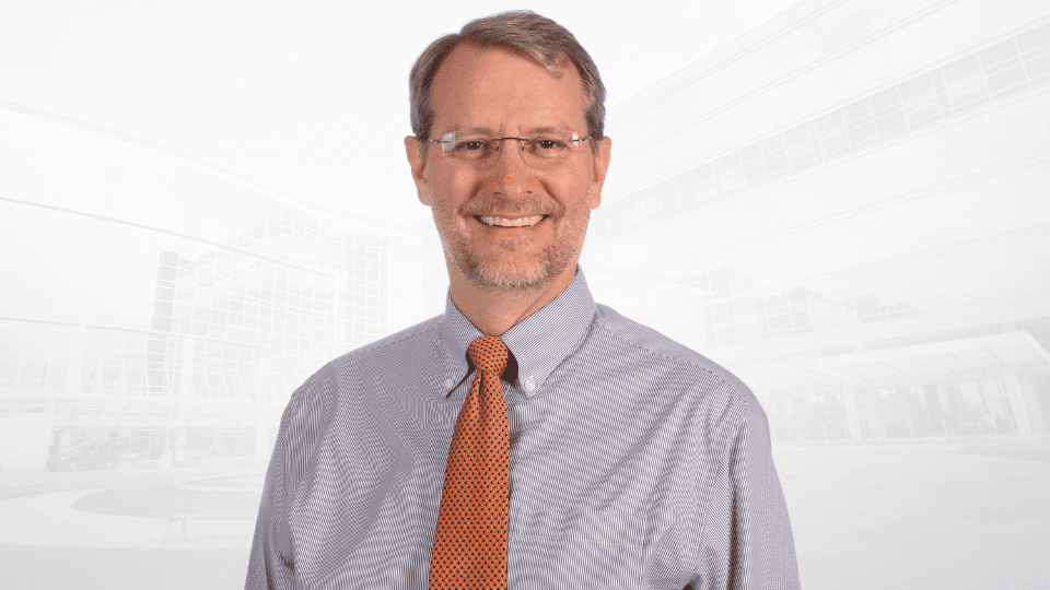 Headshot of Micah Hester wearing glasses, a striped shirt, and an orange tie.