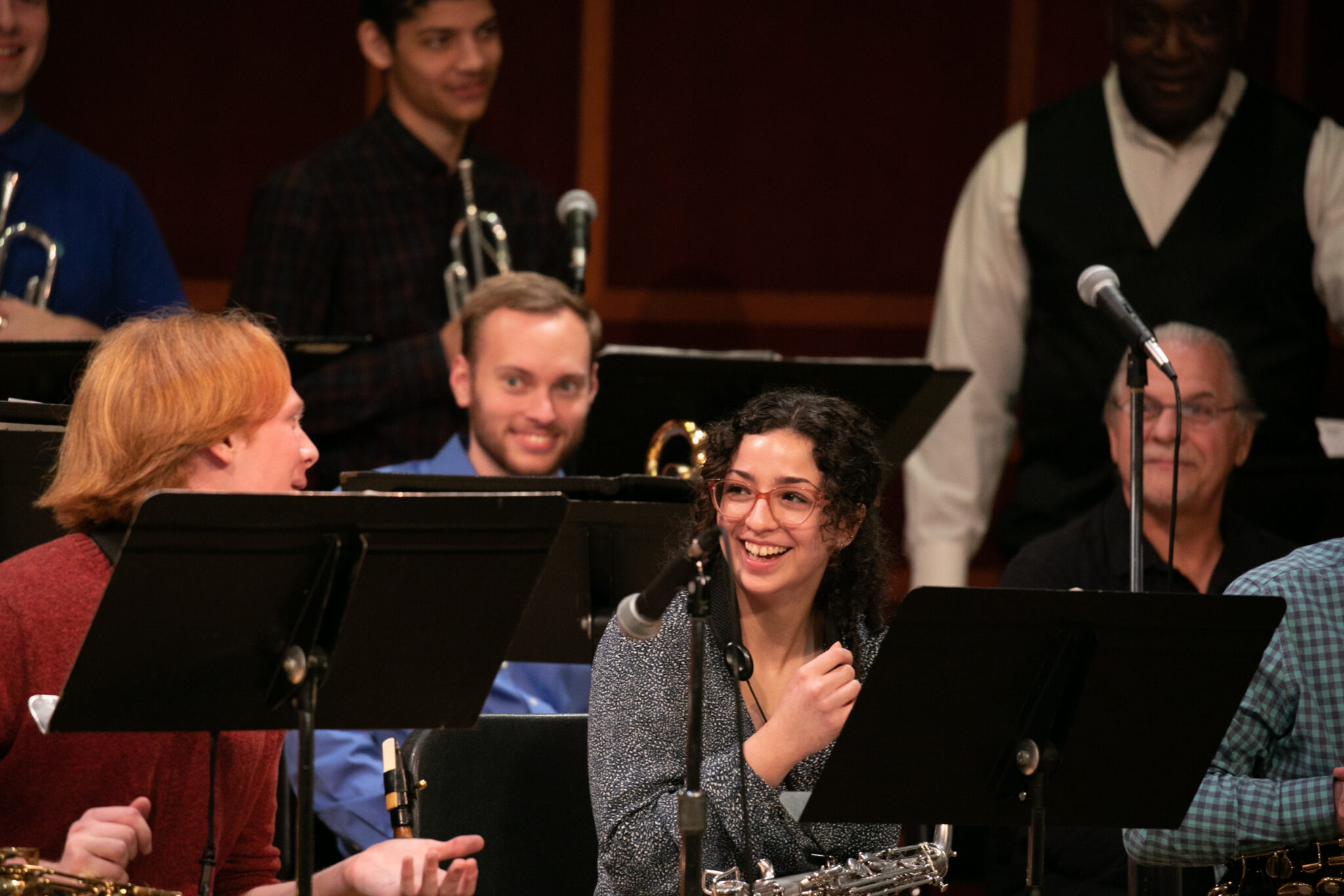 Group of musicians with various instruments, including saxophones and a trumpet, smiling and interacting behind music stands.