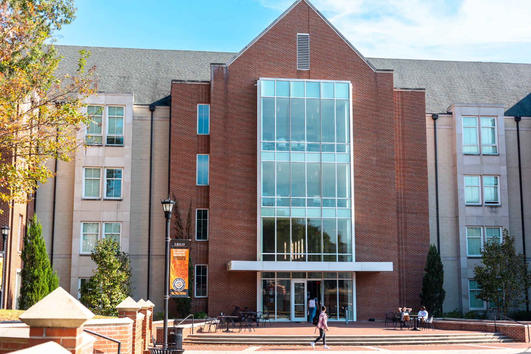 A person walking in front of a large modern building with a glass entrance on a sunny day.