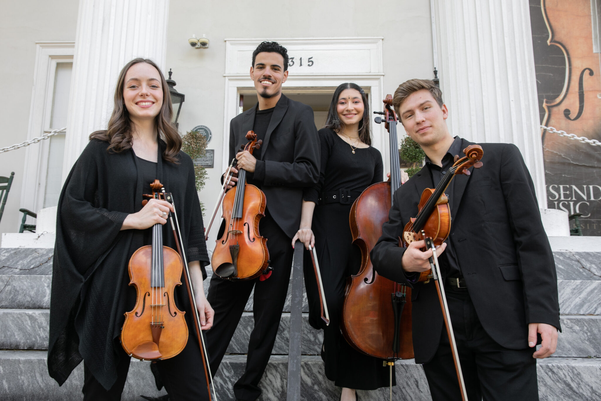 Four musicians holding string instruments pose on the steps of a building with white columns.