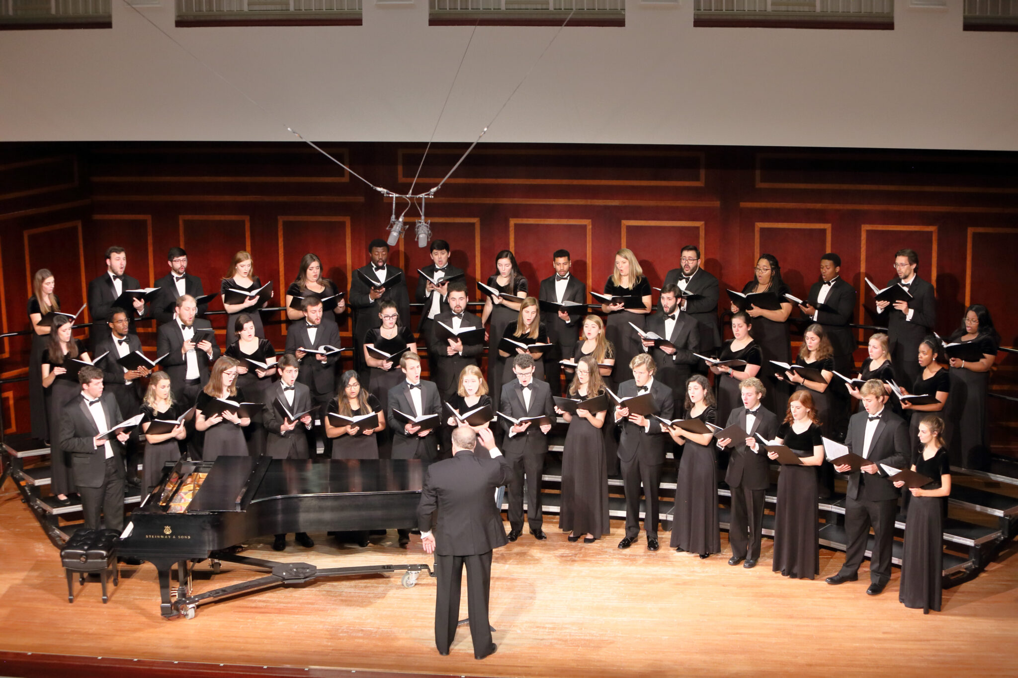 A choir performing on stage under the direction of a conductor, with a grand piano in the foreground. The choir members are wearing formal black attire.