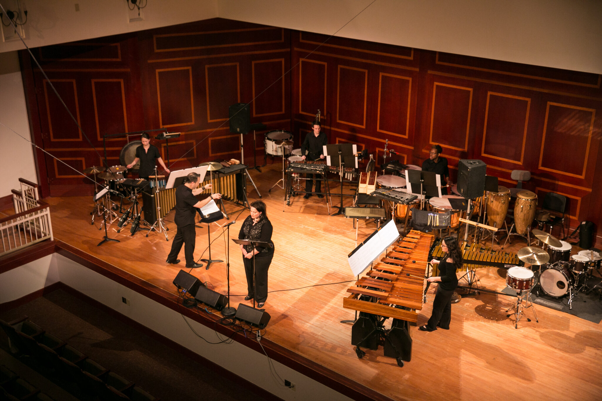 Overview of a concert hall with various percussion instruments, including marimbas and drums, set up on stage, with several musicians performing.