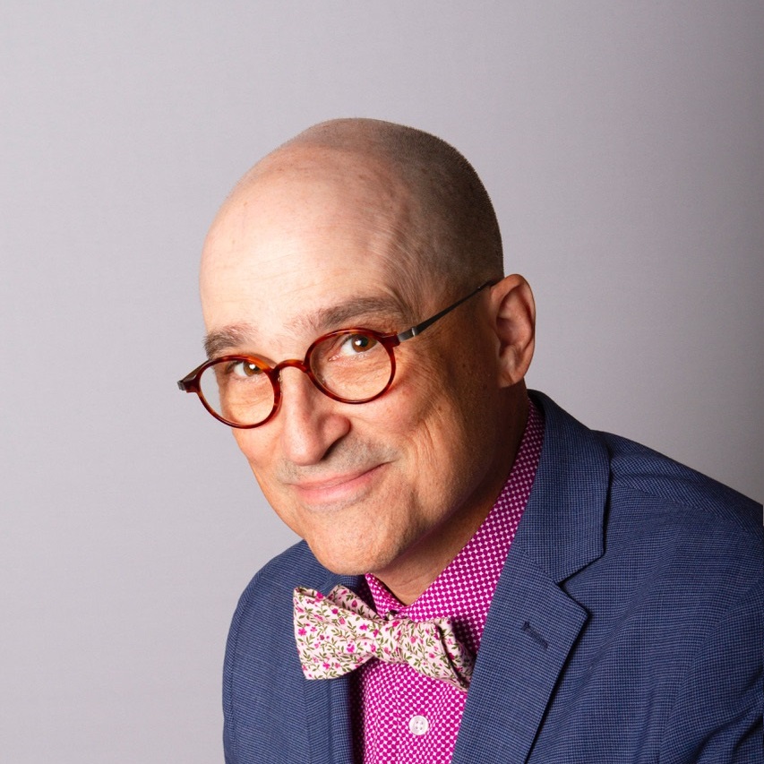 Portrait of an individual wearing round glasses, a pink patterned bow tie, and a blue blazer over a pink polka-dot shirt, smiling against a gray background.