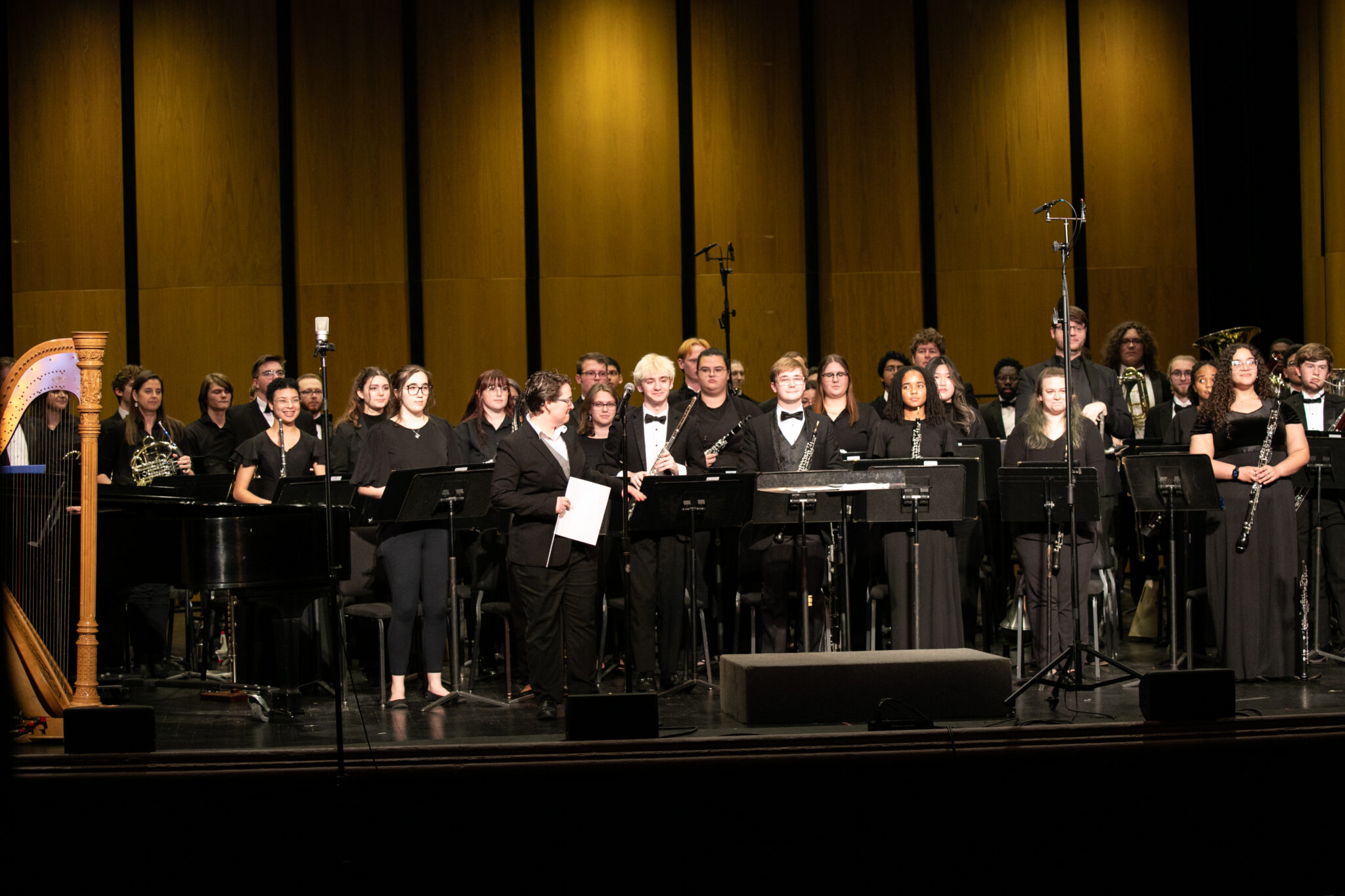 Wind ensemble on stage, featuring a variety of musicians with instruments including a harp and a piano, all dressed in formal black attire.