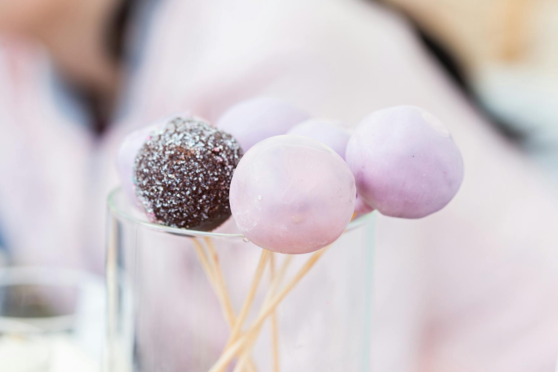 Several pink cake pops and one chocolate cake pop on sticks in a glass.