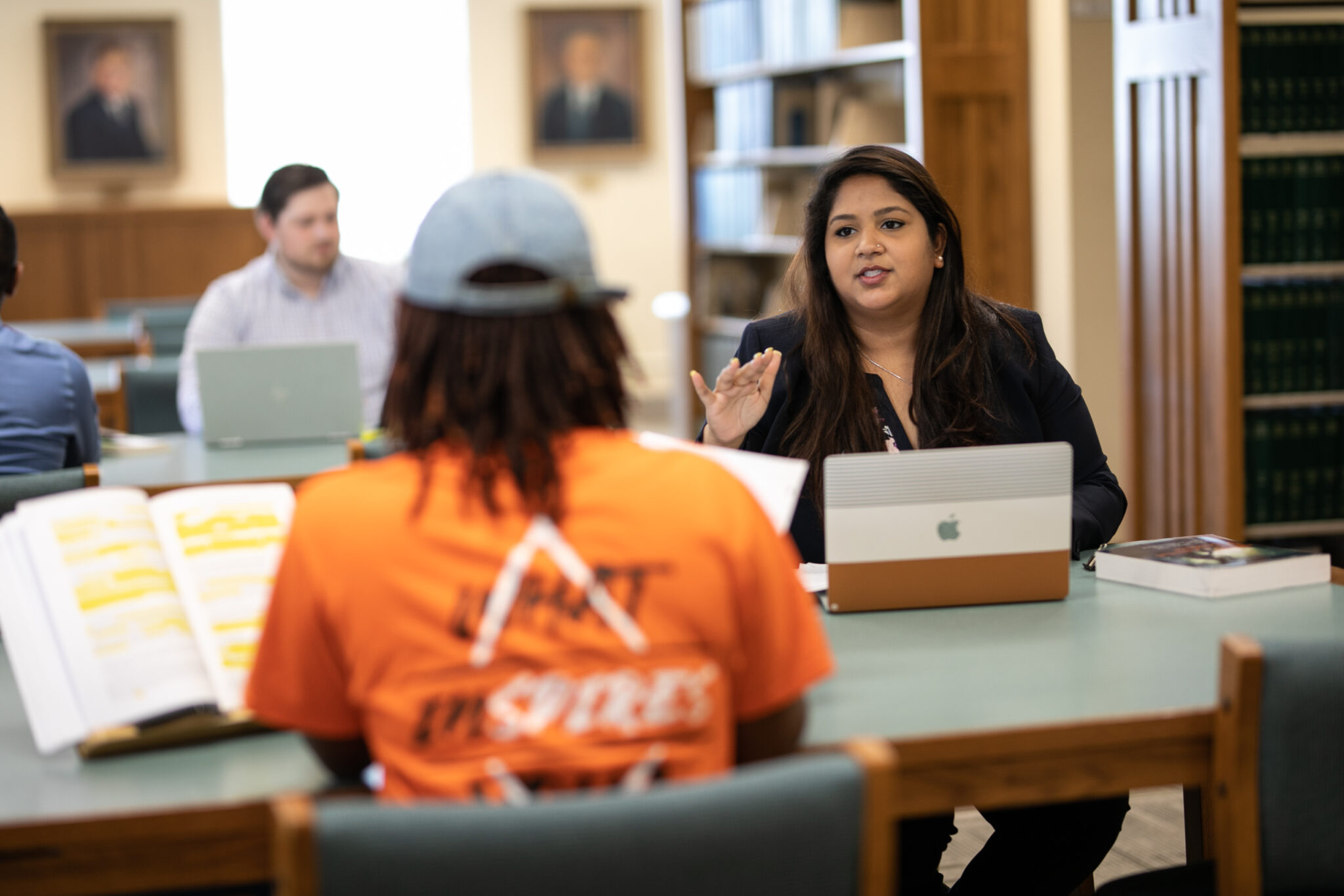 A student with a laptop in front of her talks to another student in a library.