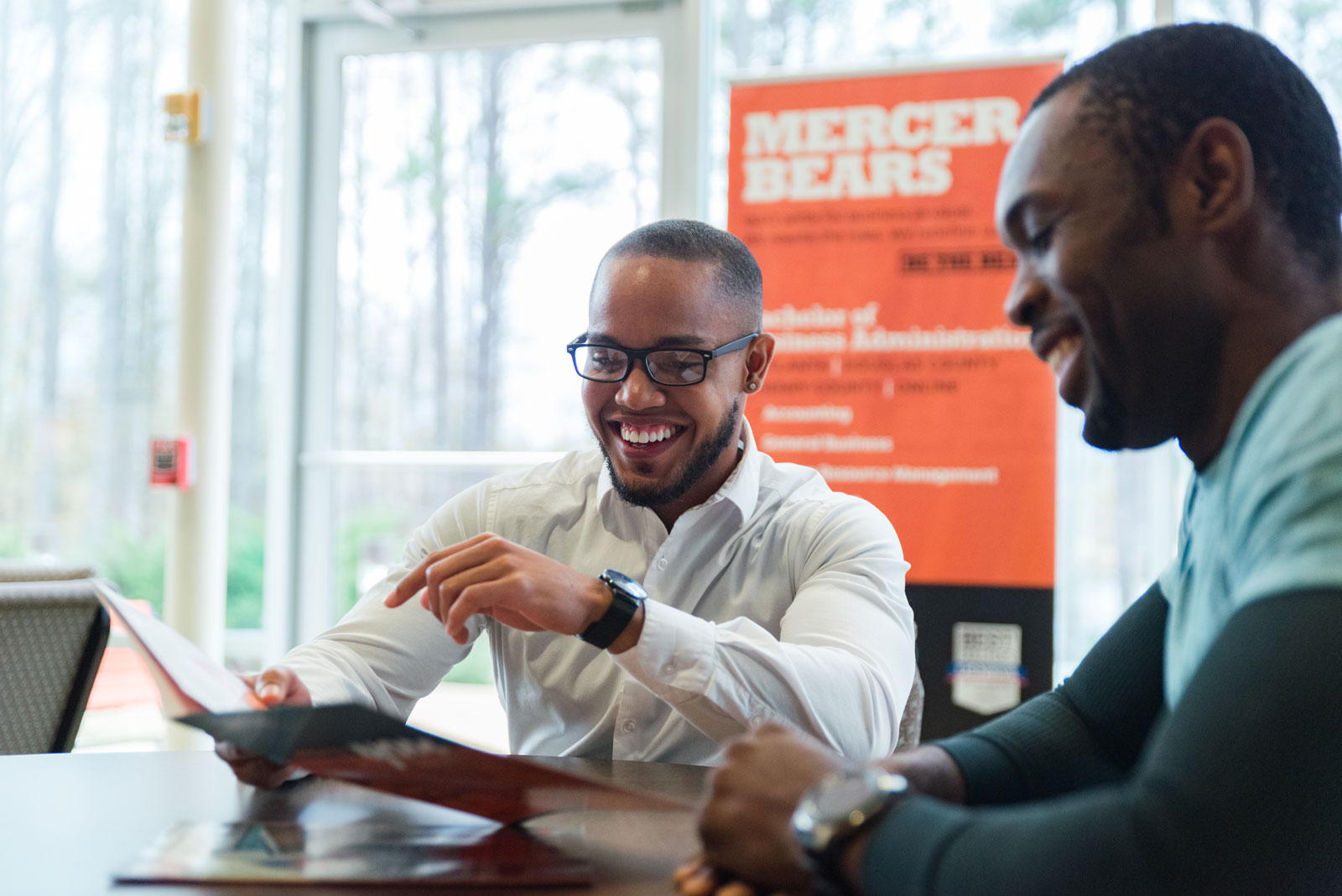 Two people are smiling and looking at an open folder at a table, with a poster reading "Mercer Bears" in the background.