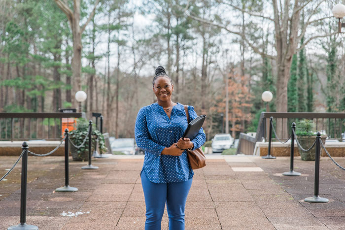 A Mercer student smiling and holding a folder stands on a walkway outdoors, surrounded by trees and lamp posts.