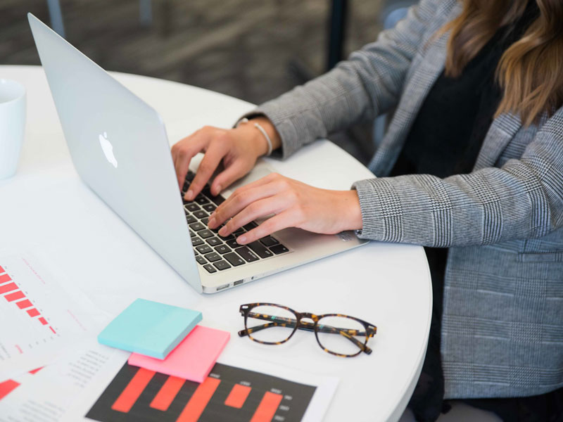 Person working on an Apple MacBook at a white table with documents, a coffee cup, and eyeglasses nearby.