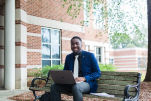 A student sits on a bench with a laptop on his lap.