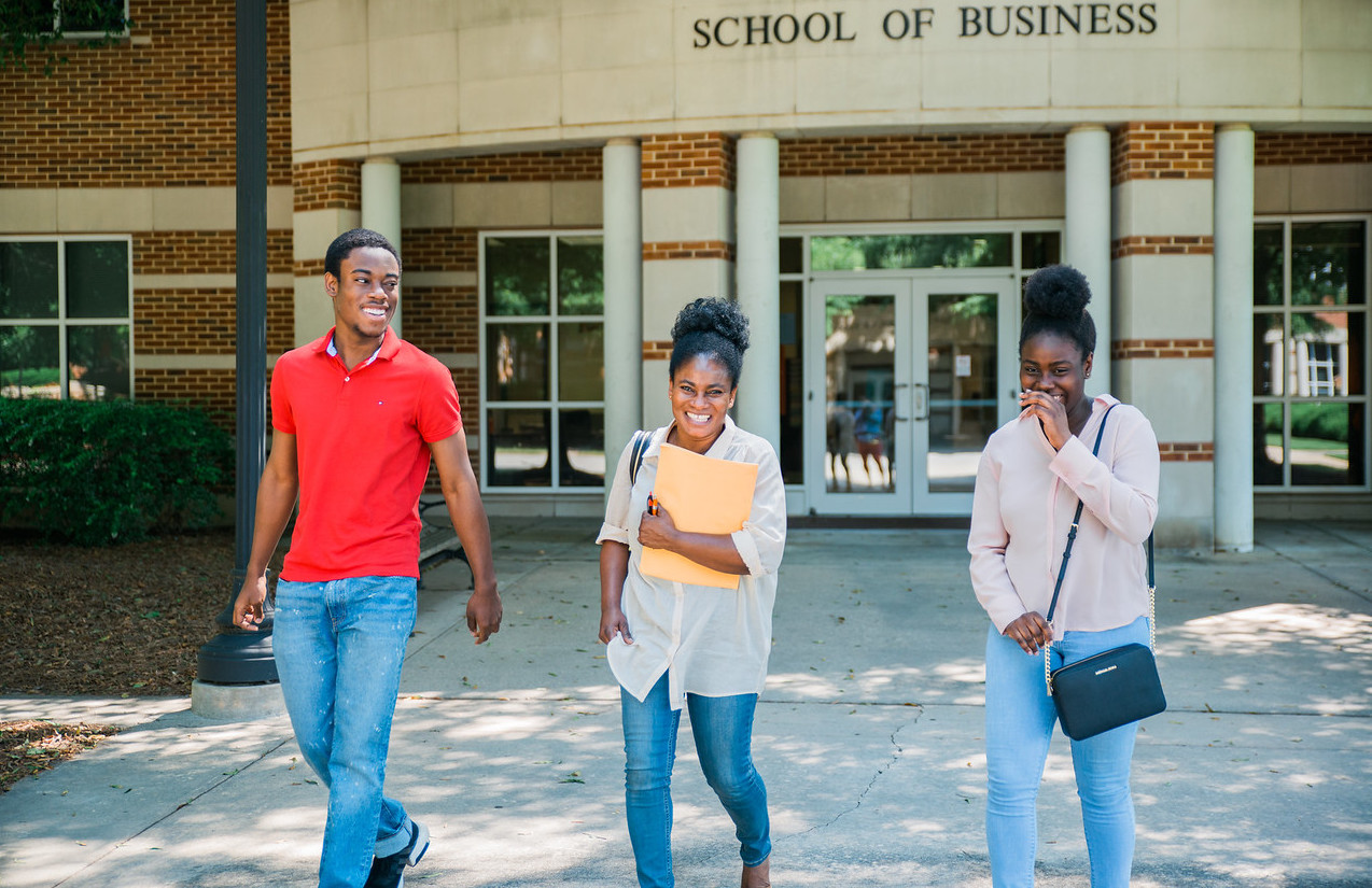 Three students walking and laughing together outside the School of Business building on a sunny day.