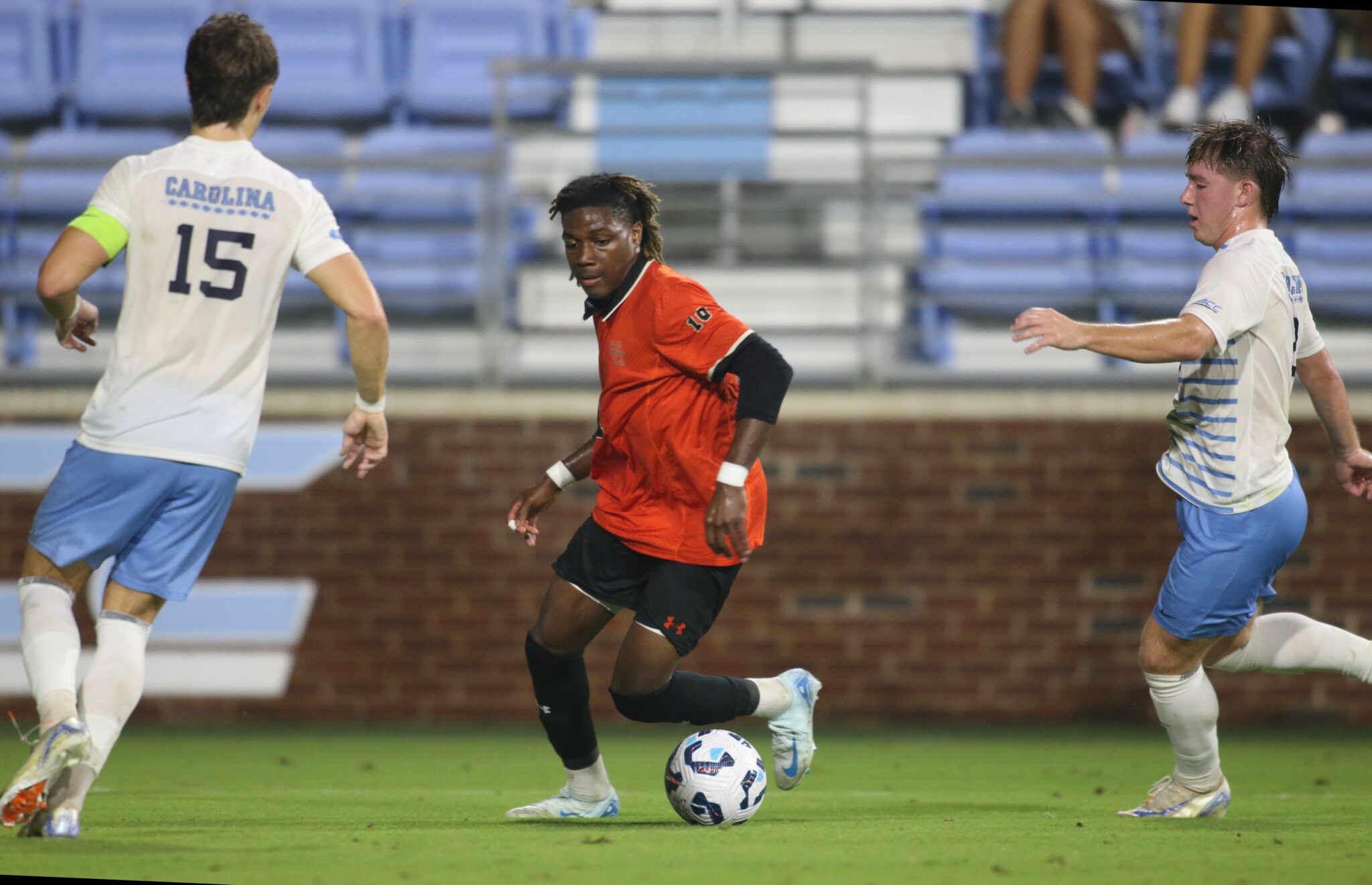 Soccer player dribbling the ball closely pursued by two opposing players during a night match.