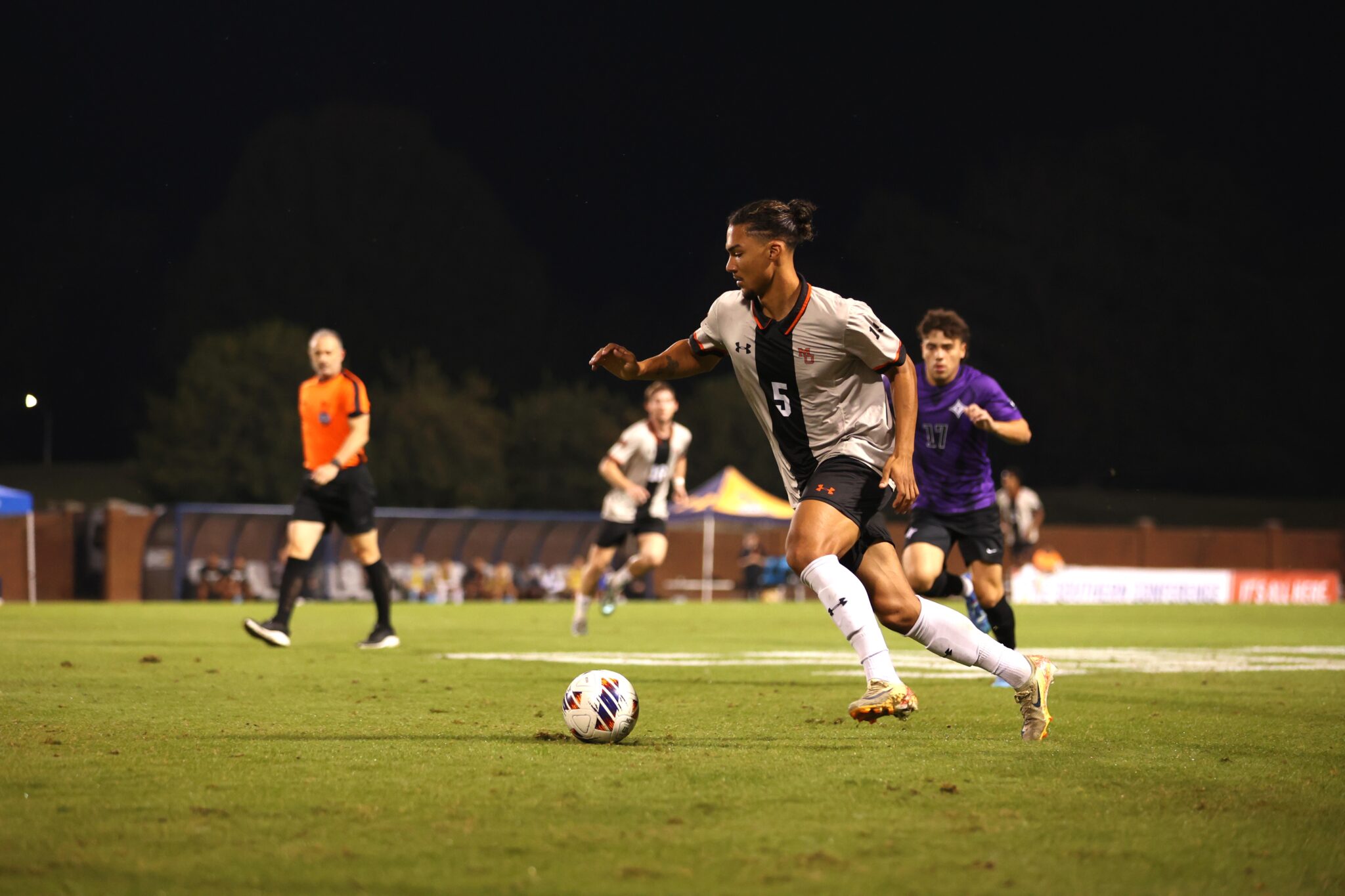 A soccer player dribbling the ball down the field during a night match, with opposing players in pursuit.