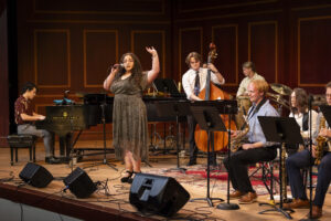 A jazz ensemble performs on stage, featuring a vocalist with a microphone, pianist, bassist and saxophonists.