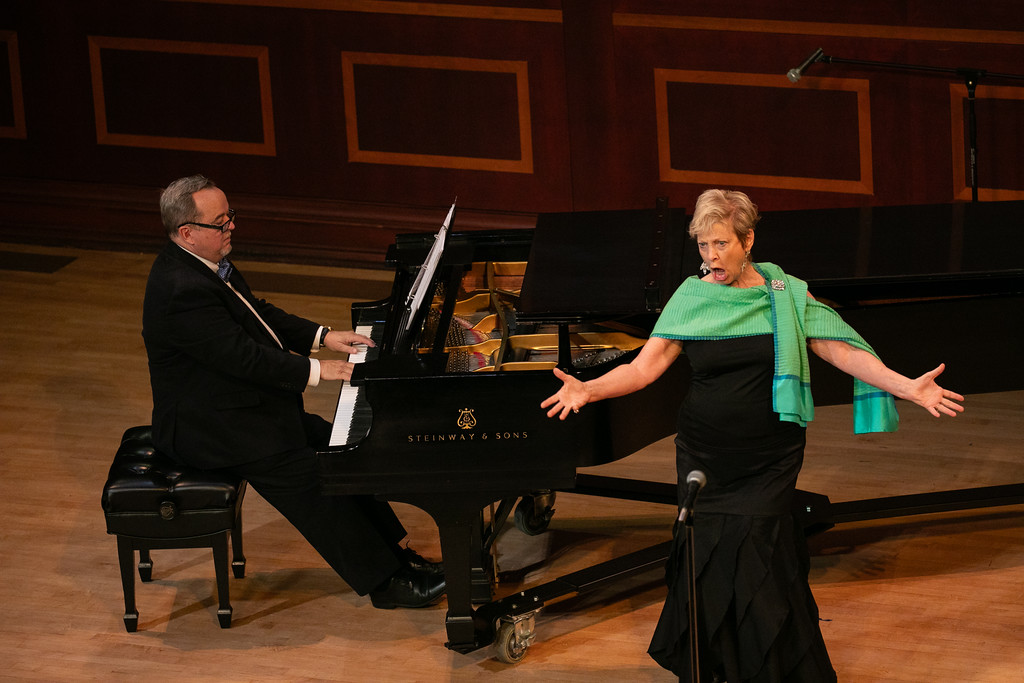 A person singing expressively with wide-open arms next to another person playing a grand piano on stage.